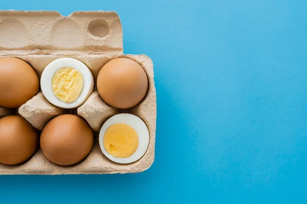 Vue du dessus des œufs cuits et crus dans un plateau en carton sur une surface bleue — Photo de stock