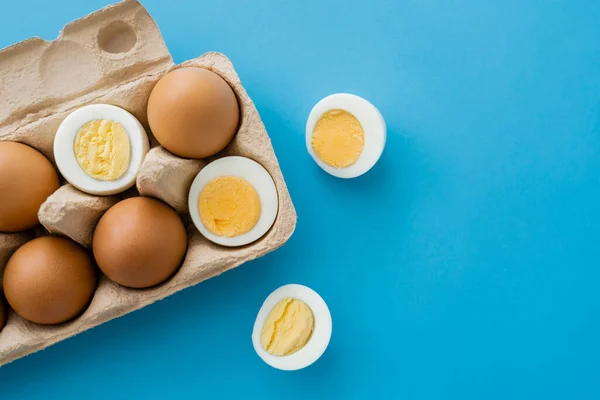 Vista superior de huevos cortados hervidos y crudos cerca de bandeja de cartón sobre fondo azul - foto de stock