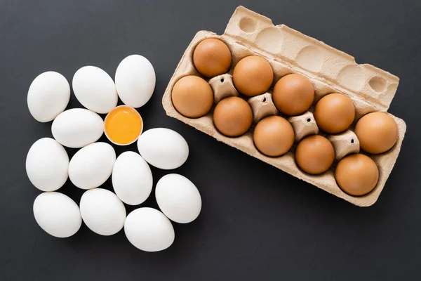Вид сверху органического желтка в скорлупе рядом с яйцами в картонной упаковке на черном фоне — стоковое фото