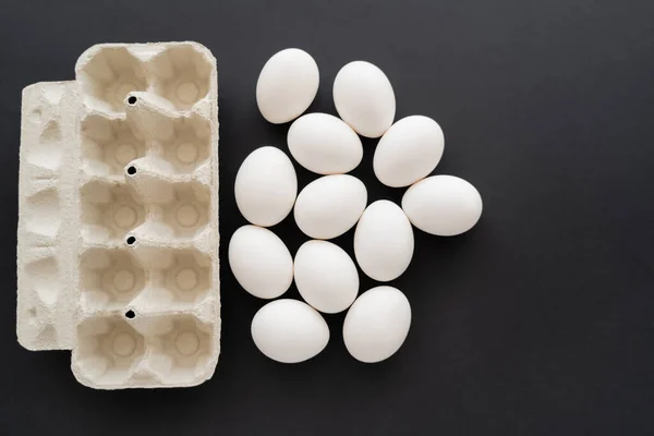 Top view of white chicken eggs near cardboard container on black background — Stock Photo