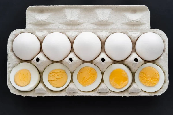 Vue du dessus des œufs de poulet crus et cuits dans un bac en carton sur fond noir — Photo de stock