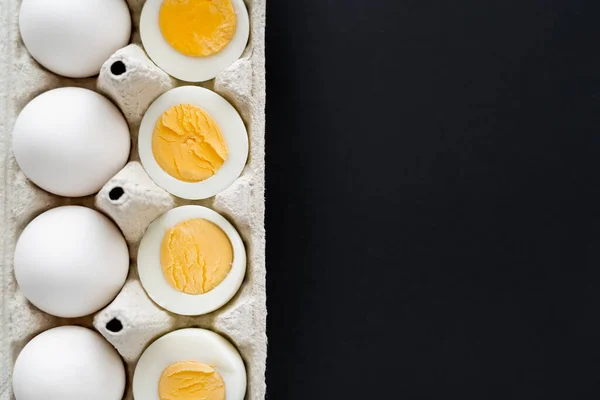 Вид сверху нарезанных вареных и сырых яиц в картонной таре, изолированной на черном фоне — стоковое фото