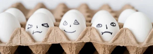 Ovos de galinha pintados com diferentes expressões faciais em bandeja de papelão sobre fundo branco, banner — Fotografia de Stock