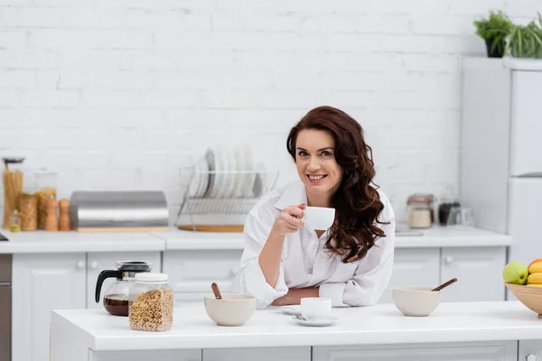 Femme souriante en chemise tenant une tasse de café près des céréales à la maison — Photo de stock