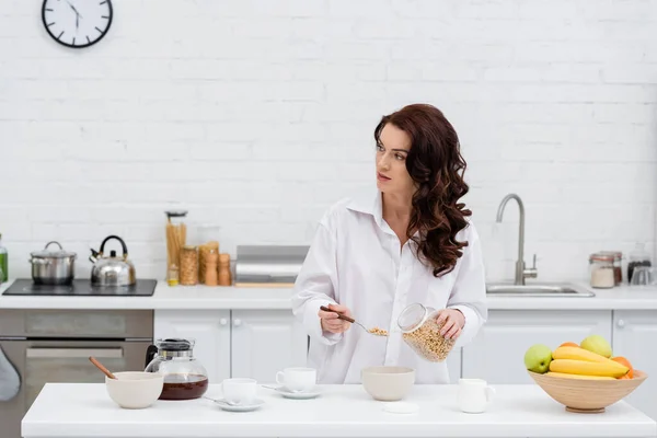 Mujer morena en camisa sosteniendo anillos de cereales cerca de café y frutas en la cocina - foto de stock