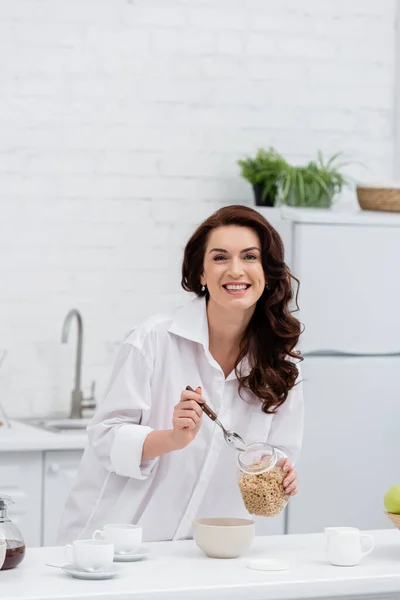 Mulher morena feliz na camisa segurando anéis de cereais perto de xícaras de café em casa — Fotografia de Stock