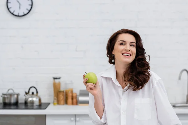 Mujer sonriente con camisa sosteniendo manzana fresca en la cocina - foto de stock