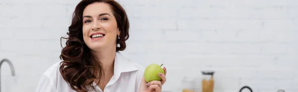 Positive brunette woman in shirt holding ripe apple in kitchen, banner — Stock Photo