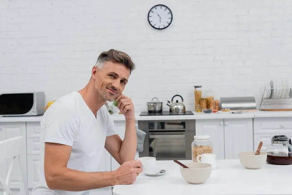 Homme souriant tenant une tasse de café et regardant la caméra près des anneaux de céréales dans la cuisine — Photo de stock