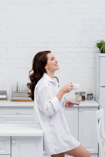 Mujer morena sonriente en camisa sosteniendo taza de café en la cocina - foto de stock