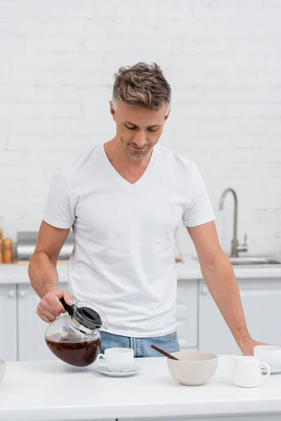 Чоловік у футболці наливає каву в чашку біля миски на столі на кухні — стокове фото