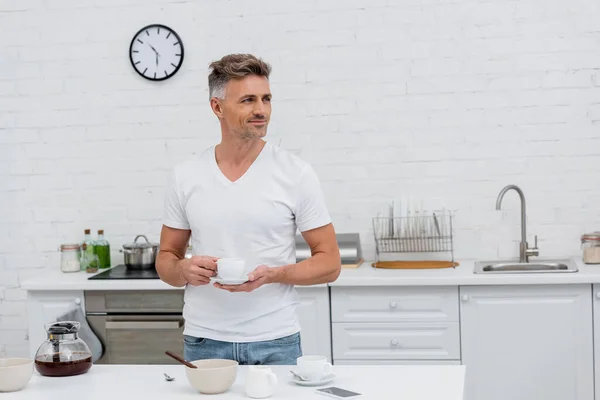 Smiling man in t-shirt holding cup of coffee near smartphone in kitchen — Stock Photo