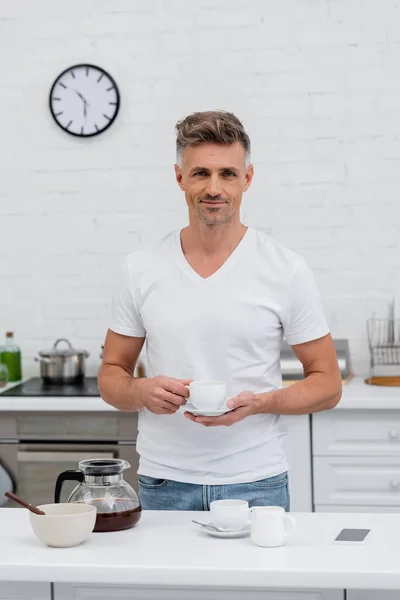 Hombre en camiseta sosteniendo taza de café cerca de teléfono inteligente con pantalla en blanco y tazón en la cocina - foto de stock