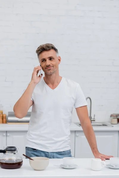 Smiling man in t-shirt talking on smartphone near coffee in kitchen — Stock Photo