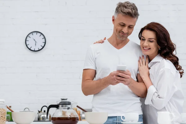 Femme positive étreignant mari avec téléphone portable près de café et petit déjeuner dans la cuisine — Photo de stock