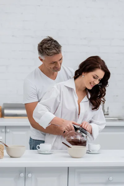 Man hugging wife in shirt pouring coffee near cereal rings in kitchen — Stock Photo
