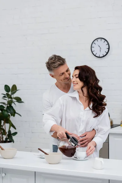 Sonriente hombre abrazando morena esposa en camisa vertiendo café en la cocina - foto de stock