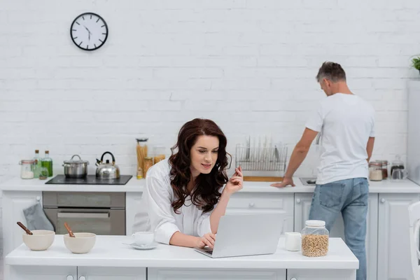 Femme brune en chemise à l'aide d'un ordinateur portable près du petit déjeuner et mari flou dans la cuisine — Photo de stock