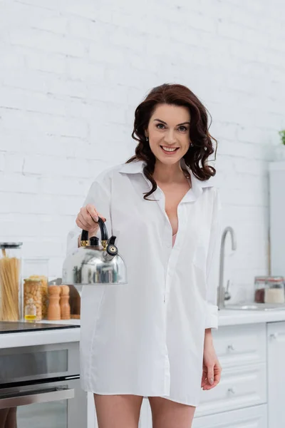 Mujer morena alegre en camisa sosteniendo hervidor de agua y mirando a la cámara en la cocina - foto de stock