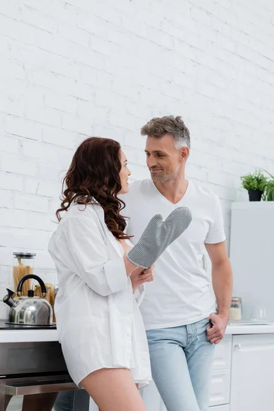 Mulher sorridente em camisa e luva de cozimento conversando com o marido na cozinha — Fotografia de Stock