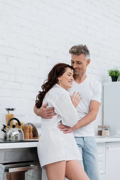 Homme souriant étreignant femme brune en chemise dans la cuisine — Photo de stock