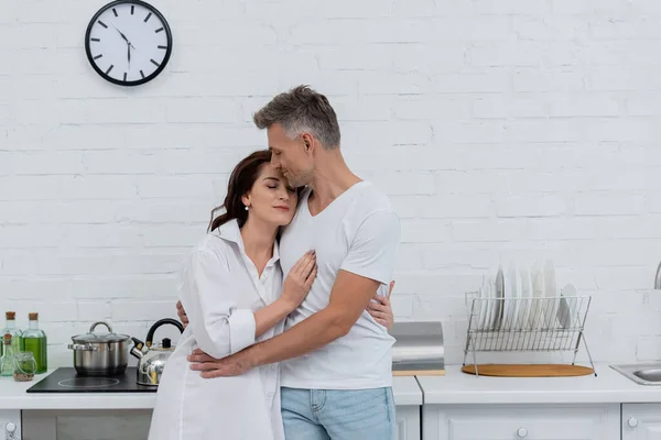Man with closed eyes hugging brunette wife in shirt in kitchen — Stock Photo