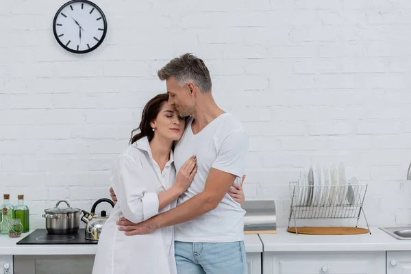 Homme aux yeux fermés étreignant femme brune en chemise dans la cuisine — Photo de stock