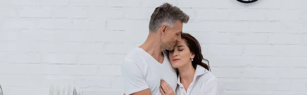 Mujer con los ojos cerrados abrazando marido en la cocina, pancarta - foto de stock