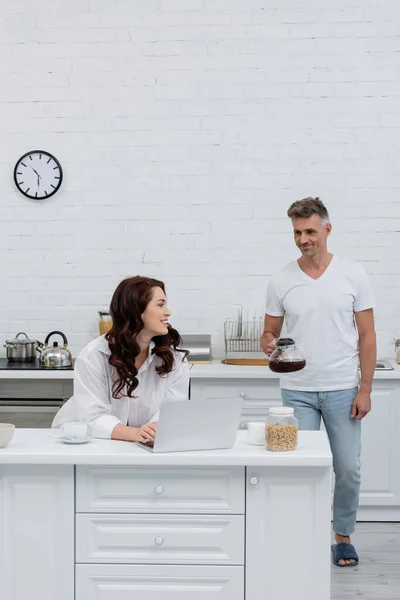 Mulher sorridente usando laptop perto do marido com panela de café e anéis de cereais na cozinha — Fotografia de Stock