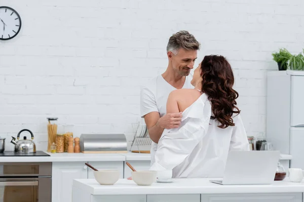 Homem positivo tirando a camisa da esposa morena perto do laptop e café na cozinha — Fotografia de Stock