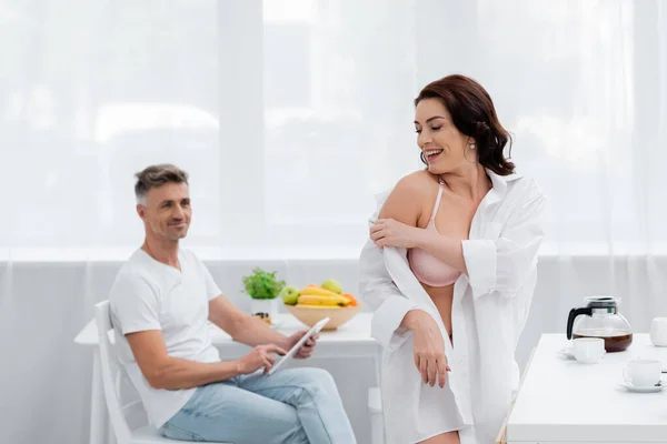 Smiling woman in bra taking off shirt near coffee and blurred husband with digital tablet in kitchen — Stock Photo