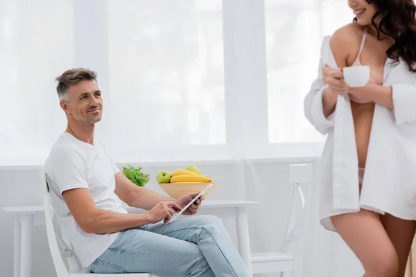 Улыбающийся мужчина, использующий цифровой планшет рядом с размытой женой в рубашке, держащий кофе на кухне — стоковое фото