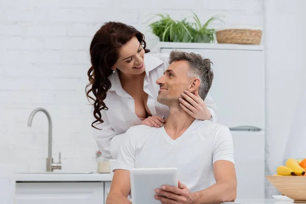 Smiling woman in shirt hugging husband with digital tablet near fruits in kitchen — Stock Photo