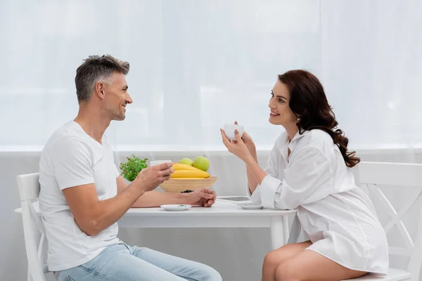 Mujer sonriente en camisa sosteniendo taza de café cerca de marido y frutas en la cocina - foto de stock