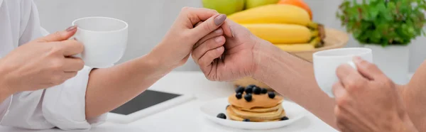 Cropped view of couple with cups of coffee holding hands near breakfast in kitchen, banner — Stock Photo