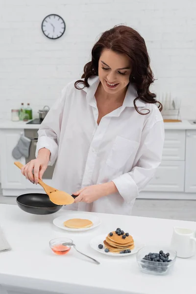 Donna sorridente in camicia che cucina frittelle vicino ai mirtilli in cucina — Foto stock