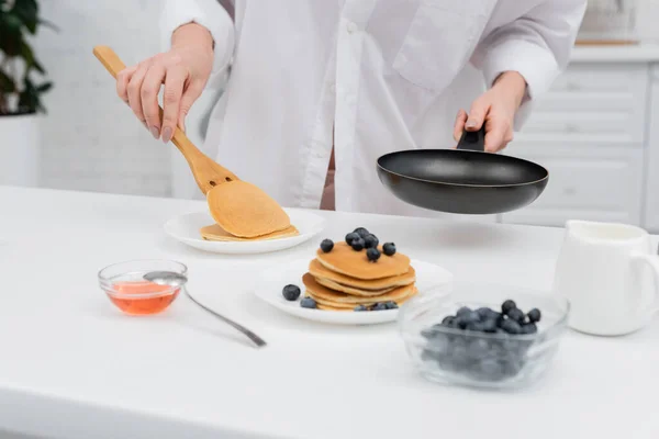 Vista ritagliata della donna in camicia che mette frittella sul piatto vicino ai mirtilli sfocati in cucina — Foto stock