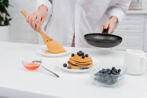 Cropped view of woman in shirt putting pancake on plate near blurred blueberries in kitchen — Stock Photo
