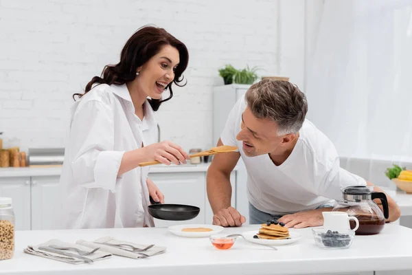 Mujer sonriente alimentando marido con panqueque cerca del desayuno en la cocina - foto de stock