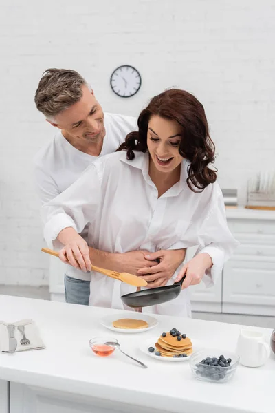 Homme étreignant femme joyeuse cuisine crêpes près des bleuets et du miel dans la cuisine — Photo de stock