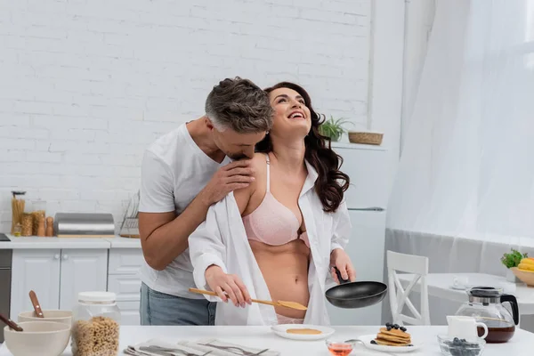 Мужчина целует плечо сексуальной жены в рубашке и нижнем белье возле завтрака на кухне — стоковое фото