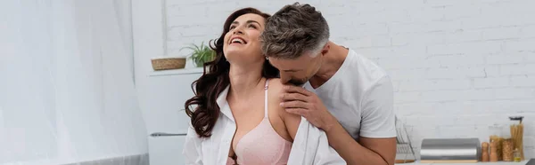 Uomo baciare spalla di moglie sorridente in reggiseno e camicia in cucina, banner — Foto stock