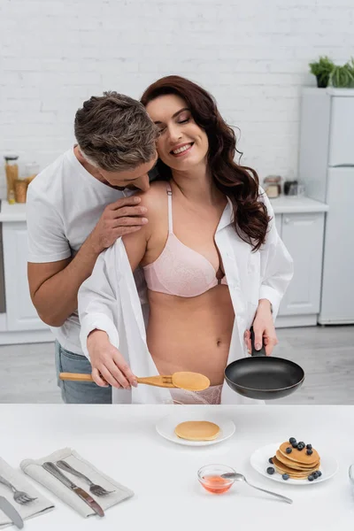 Hombre besando seductora esposa en lencería y camisa cerca de panqueques en cocina - foto de stock