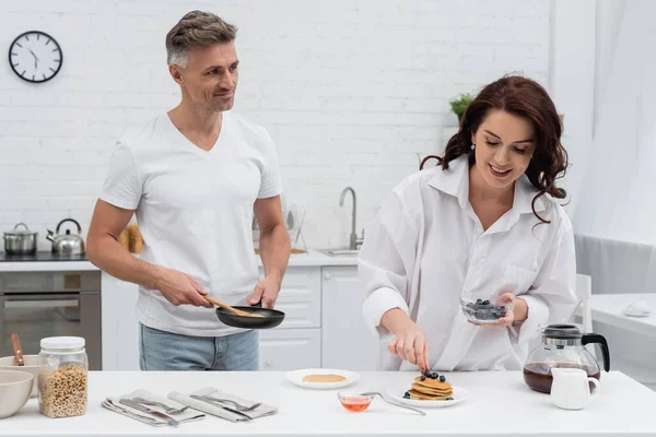 Sorrindo homem segurando frigideira perto da esposa colocando mirtilos em panquecas na cozinha — Fotografia de Stock