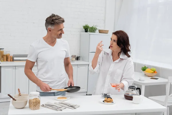 Woman in shirt talking to husband with frying pan near breakfast and coffee in kitchen — Stock Photo