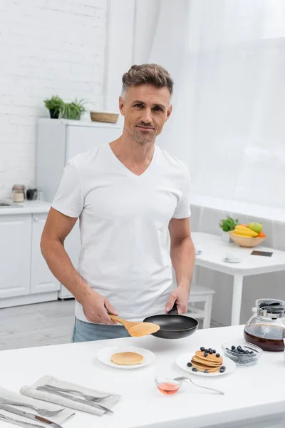 Smiling man holding frying pan near pancakes and blueberries in kitchen — Stock Photo