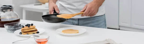 Vista cortada do homem segurando panqueca e frigideira perto de café e mirtilos na cozinha, banner — Fotografia de Stock