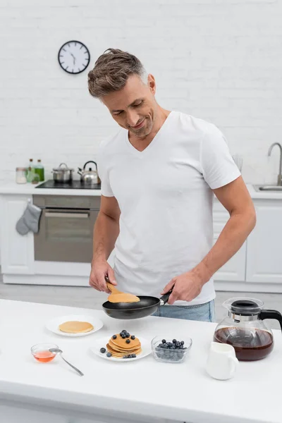 Homem positivo segurando frigideira perto de panquecas saborosas com mirtilos na cozinha — Fotografia de Stock