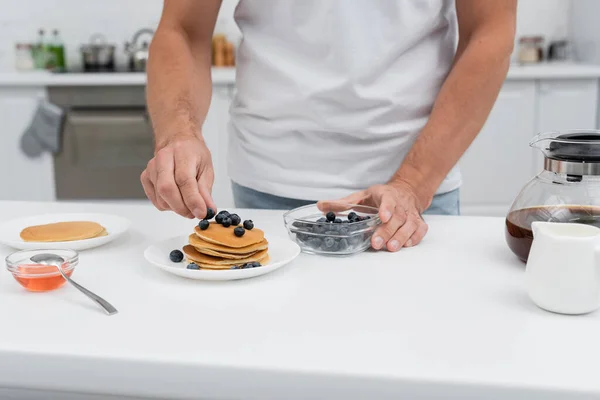 Vista recortada del hombre poniendo arándanos en panqueques cerca de la cafetera en la cocina - foto de stock