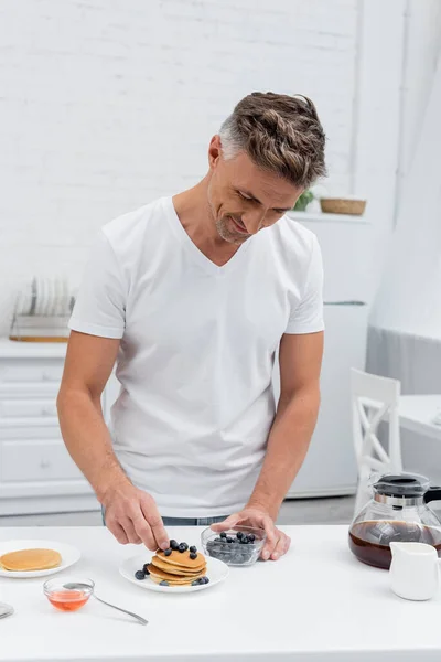 Man putting fresh blueberries on pancakes near coffee and honey in kitchen — Stock Photo